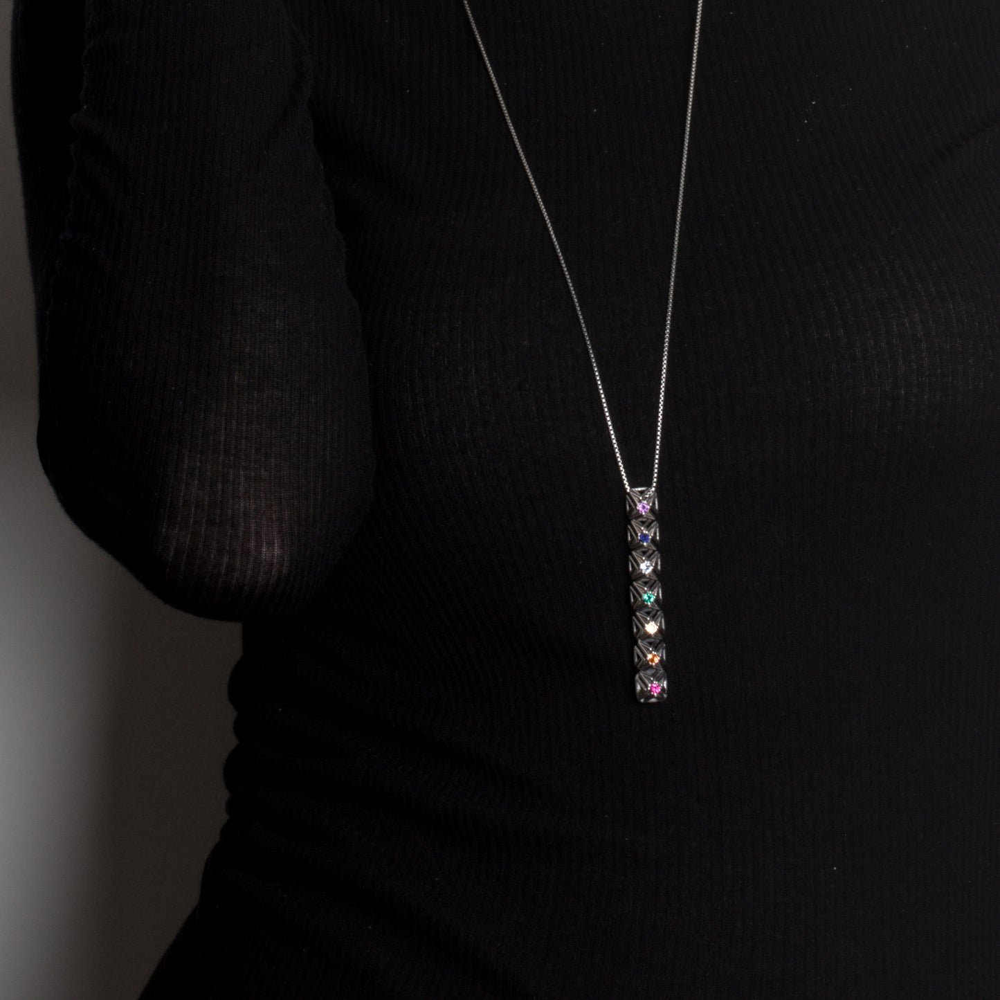 Close up of .925 sterling silver studded bar pendant set with colorful faceted gemstones. The necklace's detailed lattice stands out in edgy contrast against the black designer top it's worn with.