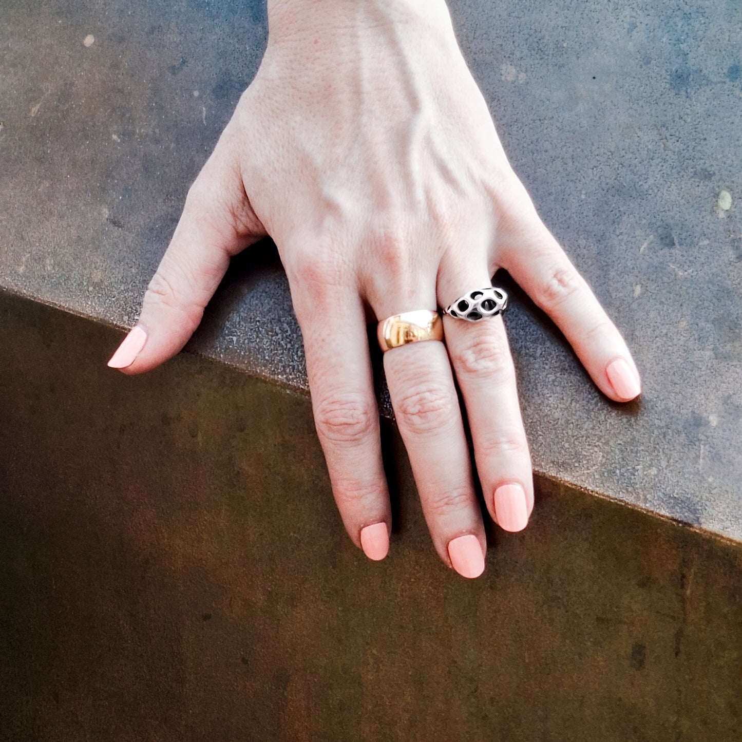 Hand wearing a sterling silver sculptural ring with puffed lattice crown next to a gold band against an edgy abstract background.