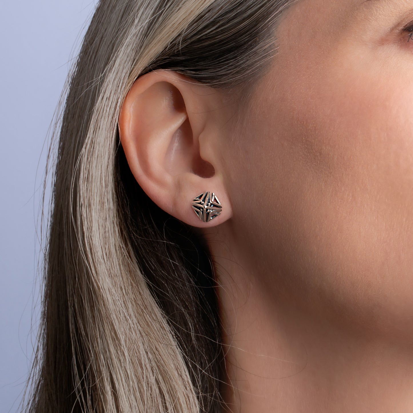 Close-up of an ear wearing a silver geometric pyramid stud earring against a light blue background.