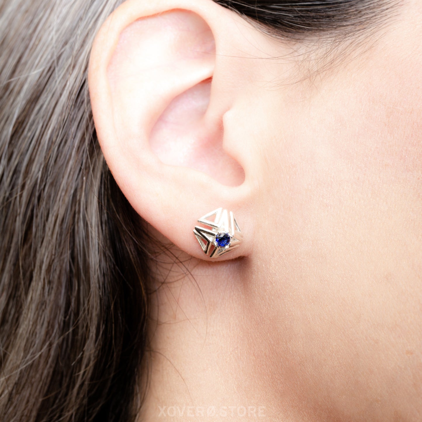 Close up of a sterling silver latticework stud earring hand-set with a lab-grown faceted sapphire shown on the ear for scale.
