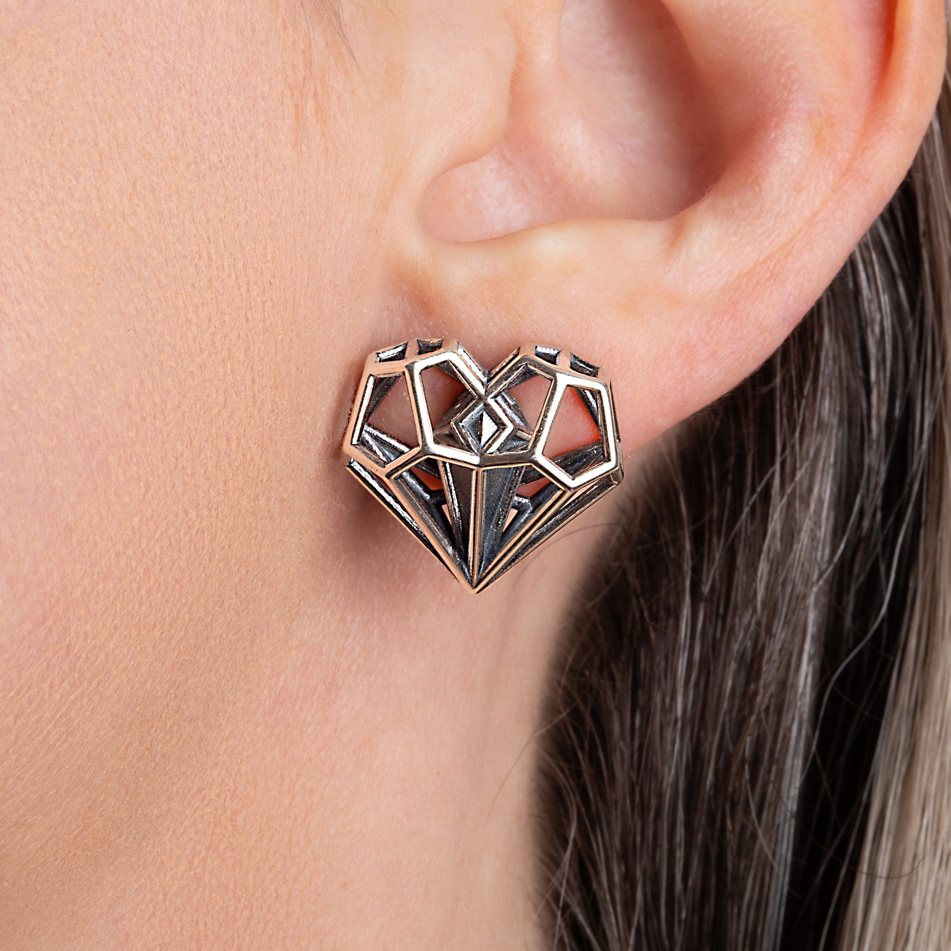 Sterling silver geometric heart-shaped earring with lattice cage and a dagger shaped solid core worn on an ear. 