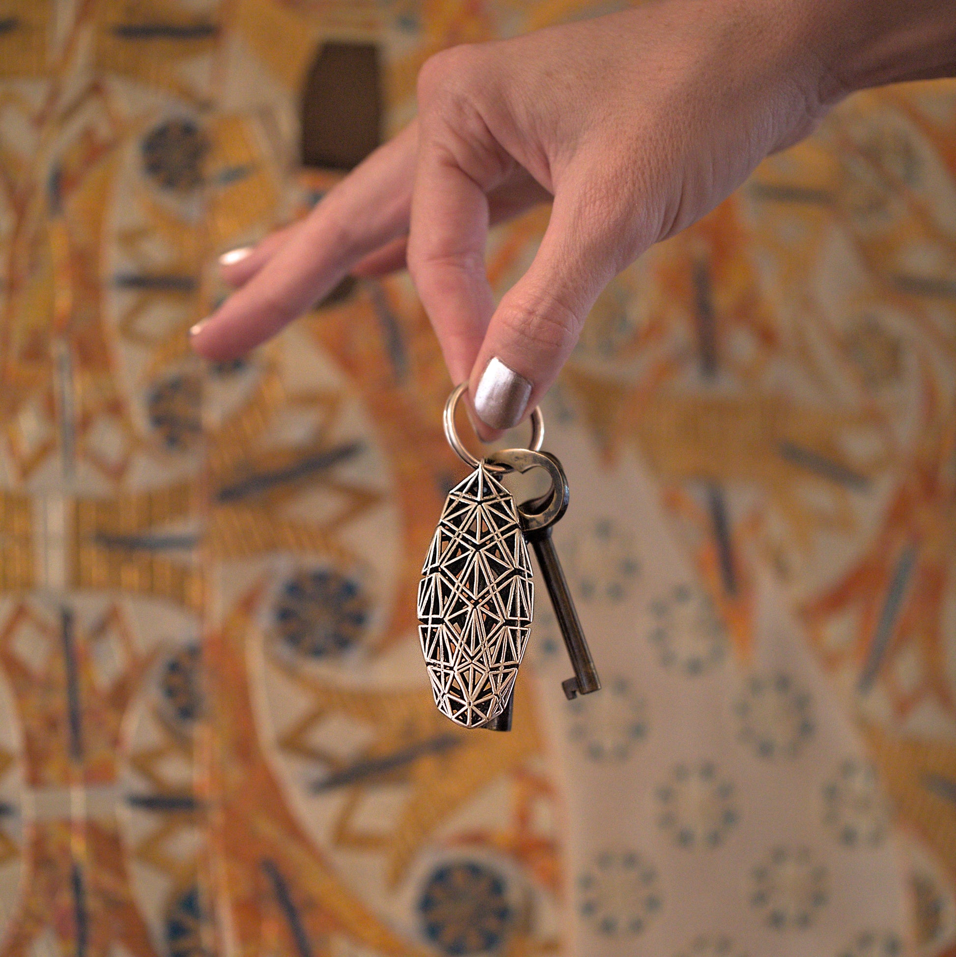 A woman's hand holding a bold sterling silver keychain defined by edgy dynamic geometry against a detailed precious metal background. 