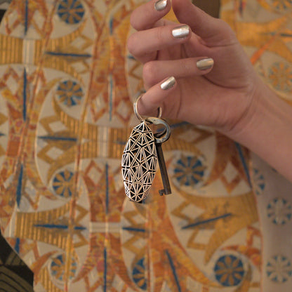 A sterling silver keychain defined by precise architectural geometry hangs from a woman's pinky finger. The key ring is large enough to accommodate multiple keys. The bold design stands out against any backdrop. 