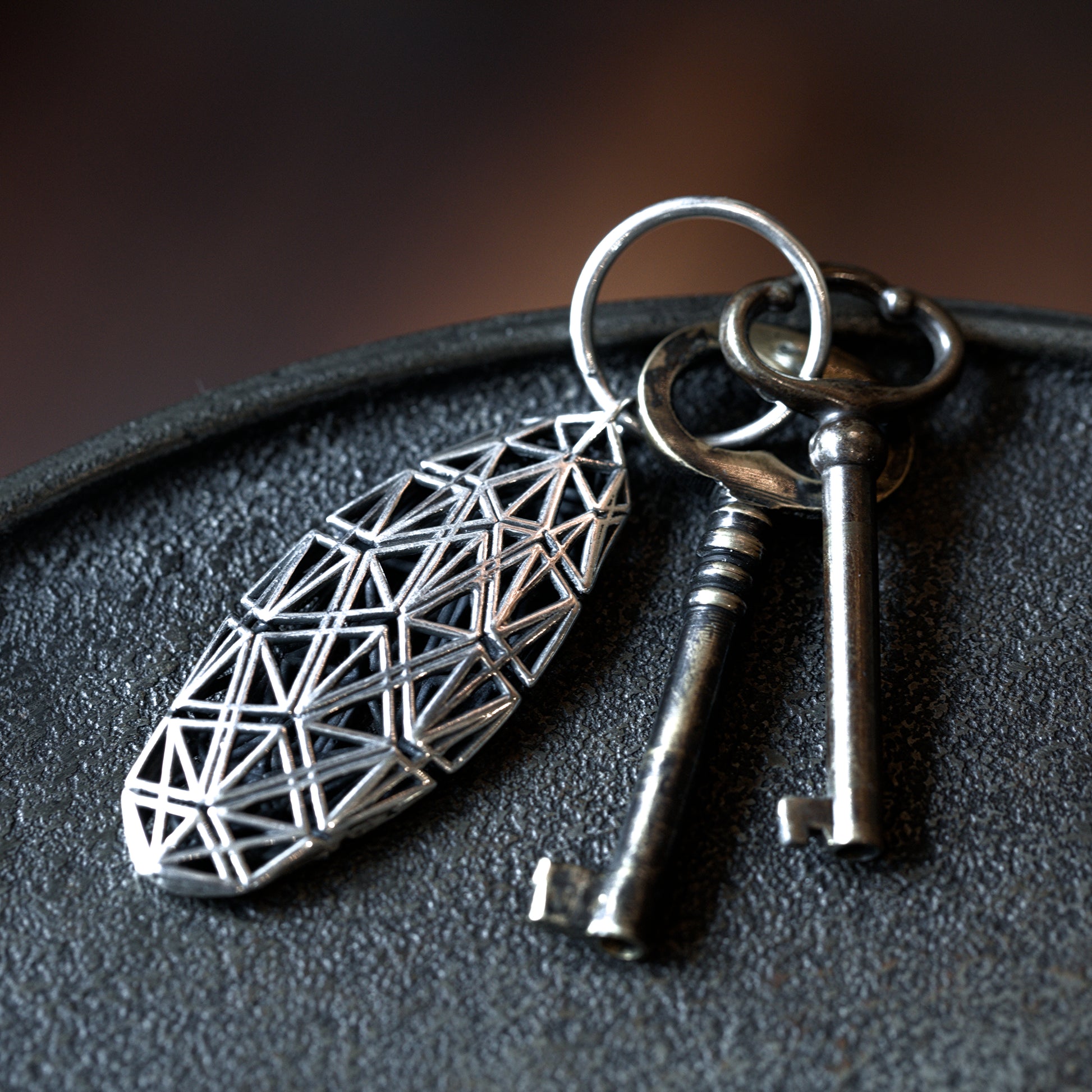 A sterling silver angular lattice designer keychain with mirror-polished surfaces, offset by contrast-oxidized matte black detailing, stands out against a dark tabletop.
