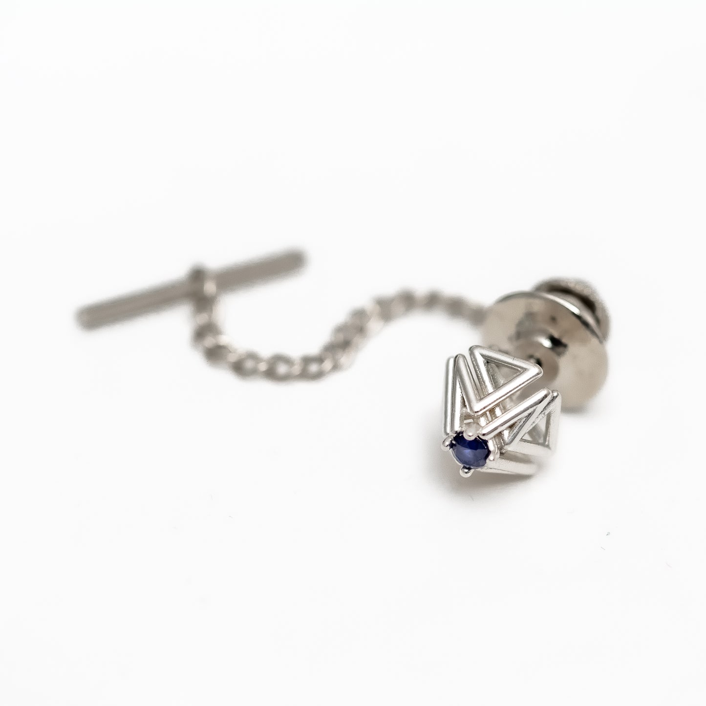 A blue sapphire and solid sterling silver tie tack with security chain rests on a white background. 
