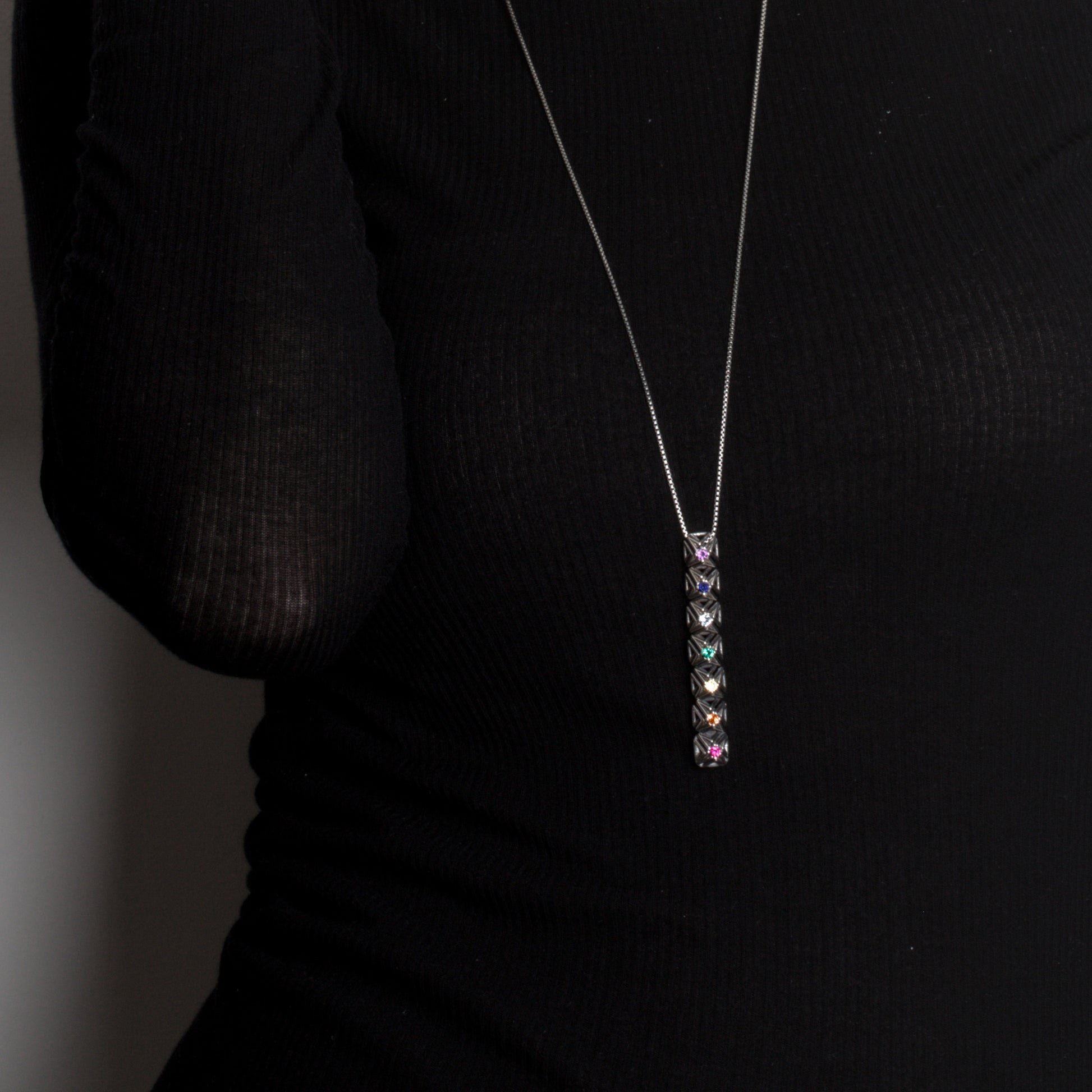 Close up of .925 sterling silver studded bar pendant set with colorful faceted gemstones. The necklace's detailed lattice stands out in edgy contrast against the black designer top it's worn with. 