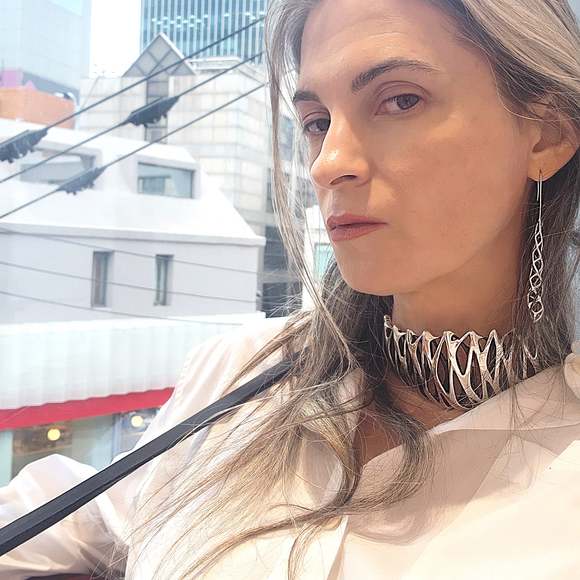 A woman wears a tall choker collar, covering most of her neck, with a crisp white button-down shirt. The solid sterling silver neckpiece is polished to a mirror shine. The necklace is secured with an X/0-embossed signature clasp, threaded through long black leather straps that drape over her shoulder.