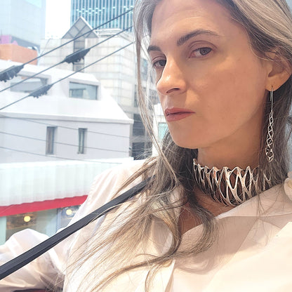 A woman wears a tall choker collar, covering most of her neck, with a crisp white button-down shirt. The solid sterling silver neckpiece is polished to a mirror shine. The necklace is secured with an X/0-embossed signature clasp, threaded through long black leather straps that drape over her shoulder.