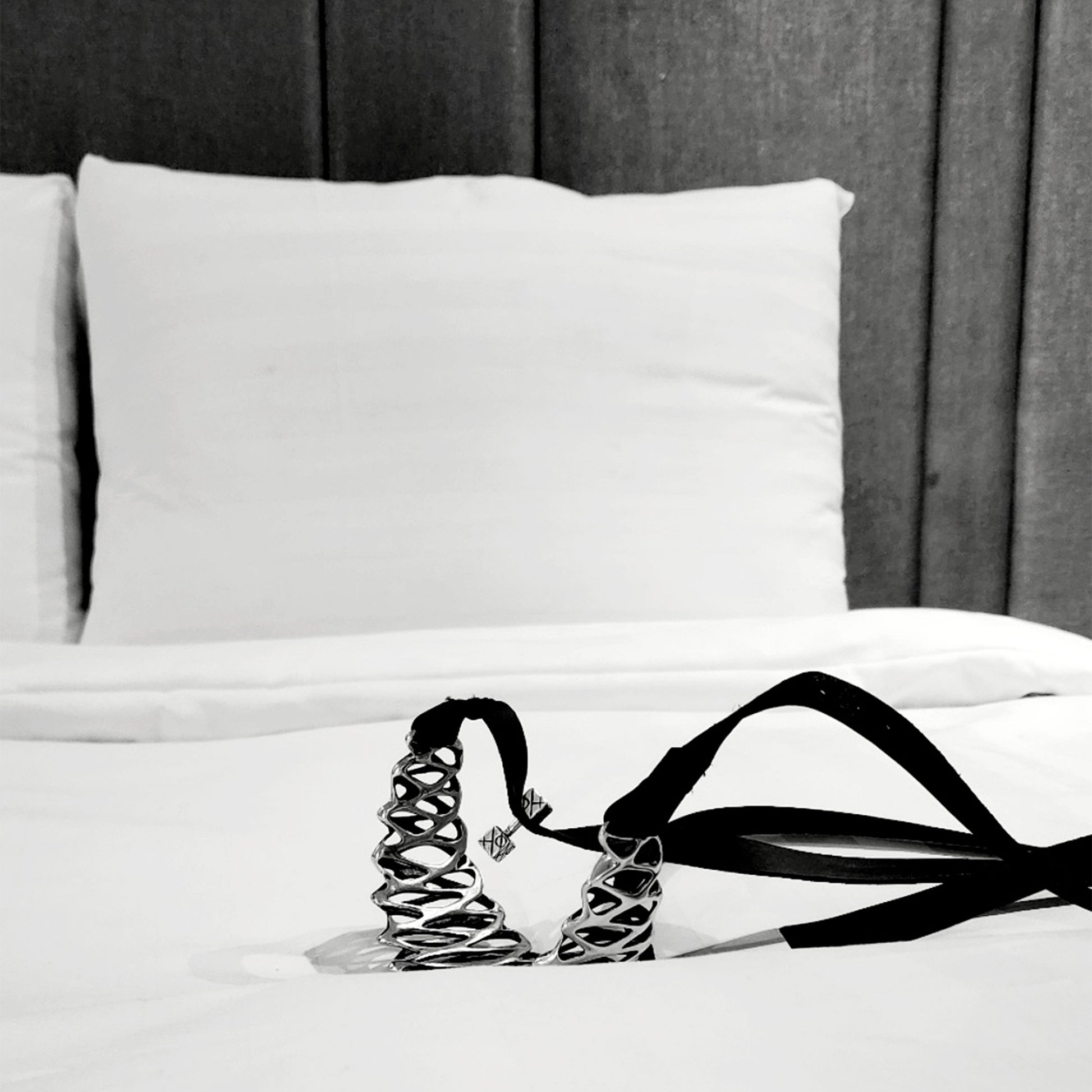 Anticipation: a mirror polished solid sterling silver statement choker necklace with long black straps rests on a freshly made bed with crisp white sheets and padded headboard. 