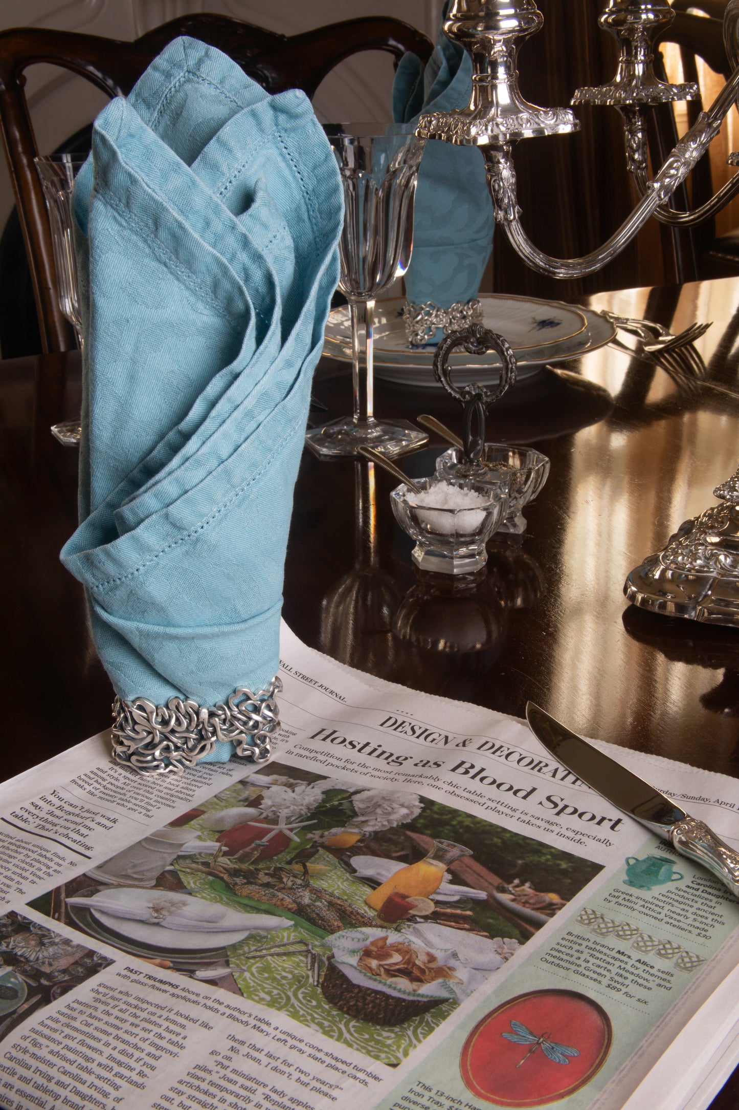 Xover0���s solid sterling silver Mississippi River napkin ring beside a Wall Street Journal article titled ���Hosting as Blood Sport,��� highlighting the competitive art of table setting.
