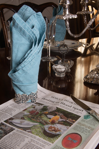 Xover0���s solid sterling silver Mississippi River napkin ring beside a Wall Street Journal article titled ���Hosting as Blood Sport,��� highlighting the competitive art of table setting.